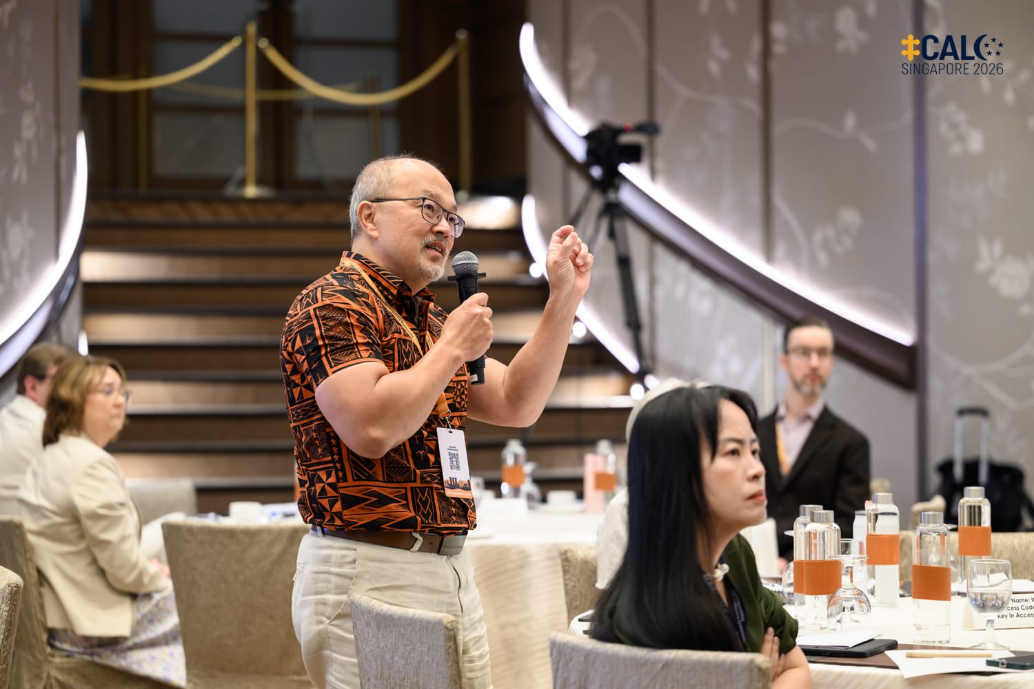 A delegate stands and speaks into a handheld microphone during a session at the Conference, with other attendees seated at round tables in the ballroom at The Fullerton Hotel Singapore.